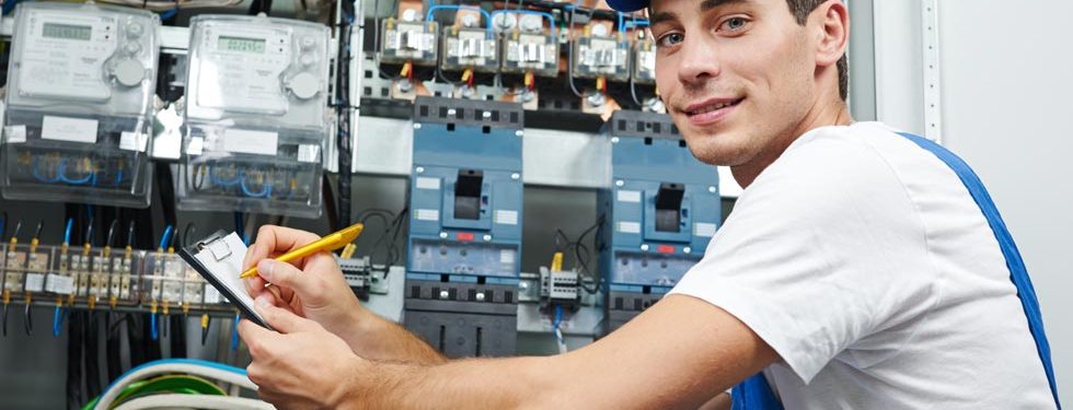 Éléctricien en train de noter sur un papier les problèmes sur le tableau électrique derrière lui